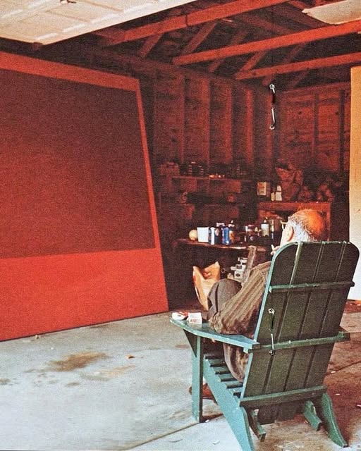 A seated figure looking at a large red painting in a timber studio