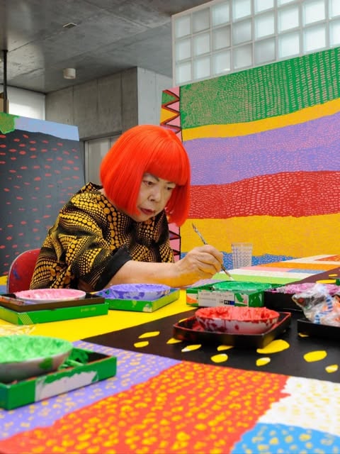 An artist at work surrounded by vividly coloured paintings and paint trays